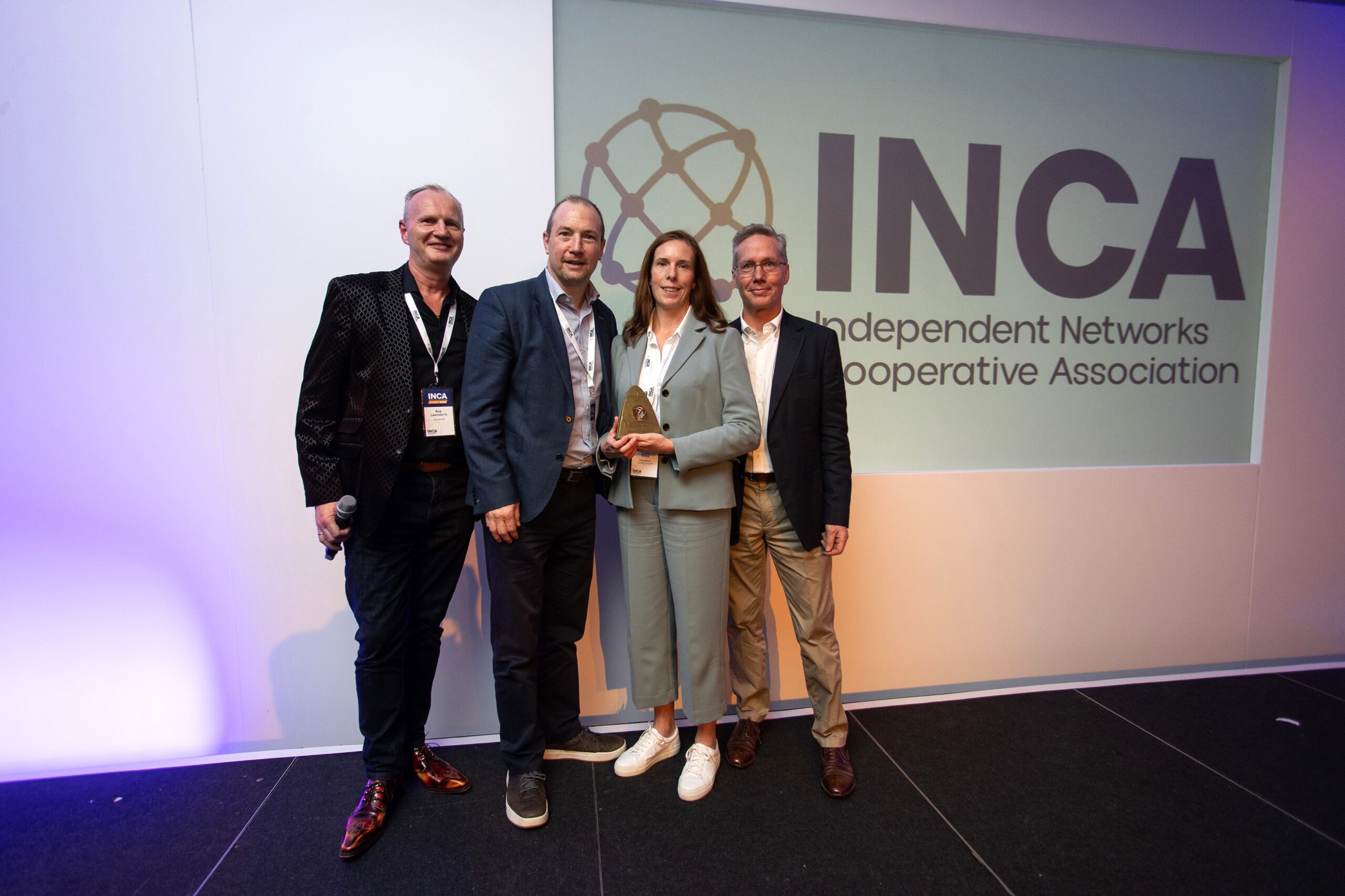 Four individuals standing on a stage in front of a screen displaying the Independent Networks Cooperative Association (INCA) logo. Anna, a member of Oxfordshire's Digital Infrastructure Programme, holds an award. The stage is lit with blue and purple lighting.
