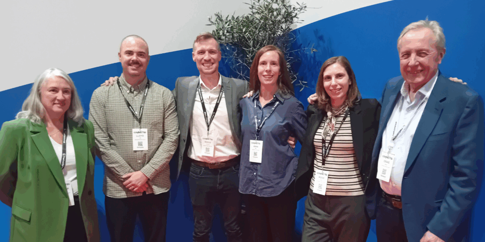 A group of six people standing together indoors against a backdrop with a curved blue design and a potted plant in the background. All individuals are wearing event lanyards with name badges, and they are dressed in business or smart-casual attire, including blazers, shirts, and trousers. 