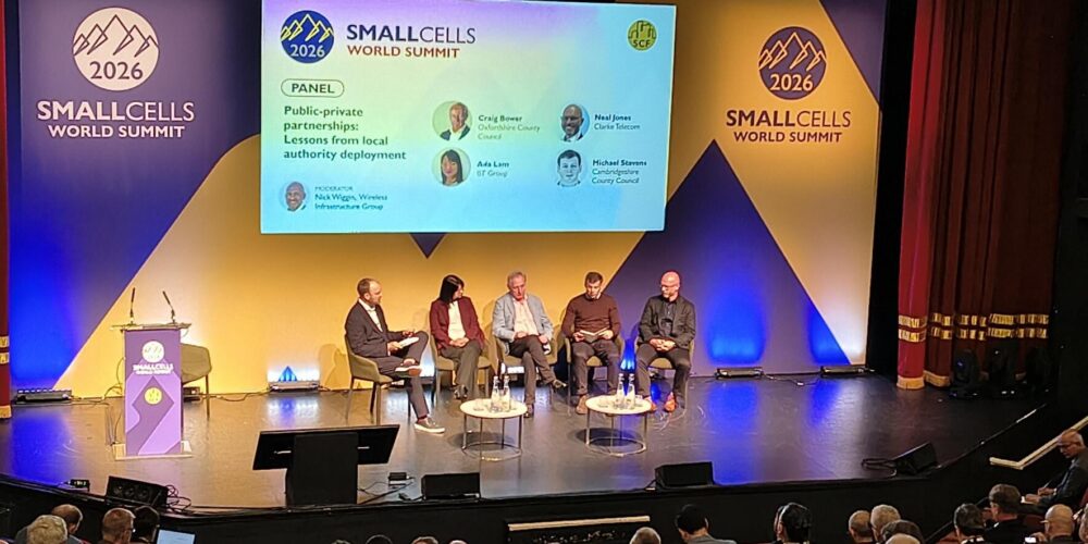 Panel discussion on stage at the Small Cells World Summit 2026, with five speakers seated in armchairs and two small round tables between them, in front of a large screen displaying the session title “Public-private partnerships: Lessons from local authority deployment.” Event branding and logos appear on both sides of the stage, while an audience is seated in rows in the foreground.