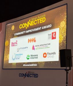 Large presentation screen at the Connected Britain Awards showing the shortlist for the Community Improvement Award. The screen displays logos of finalists including Digital Hub, Way More Than 5G, Kent County Council, Oxfordshire Digital Infrastructure Programme, Littlegarth Microbotics, Wessex Internet, Founds Group and Swansea Bay City Deal.